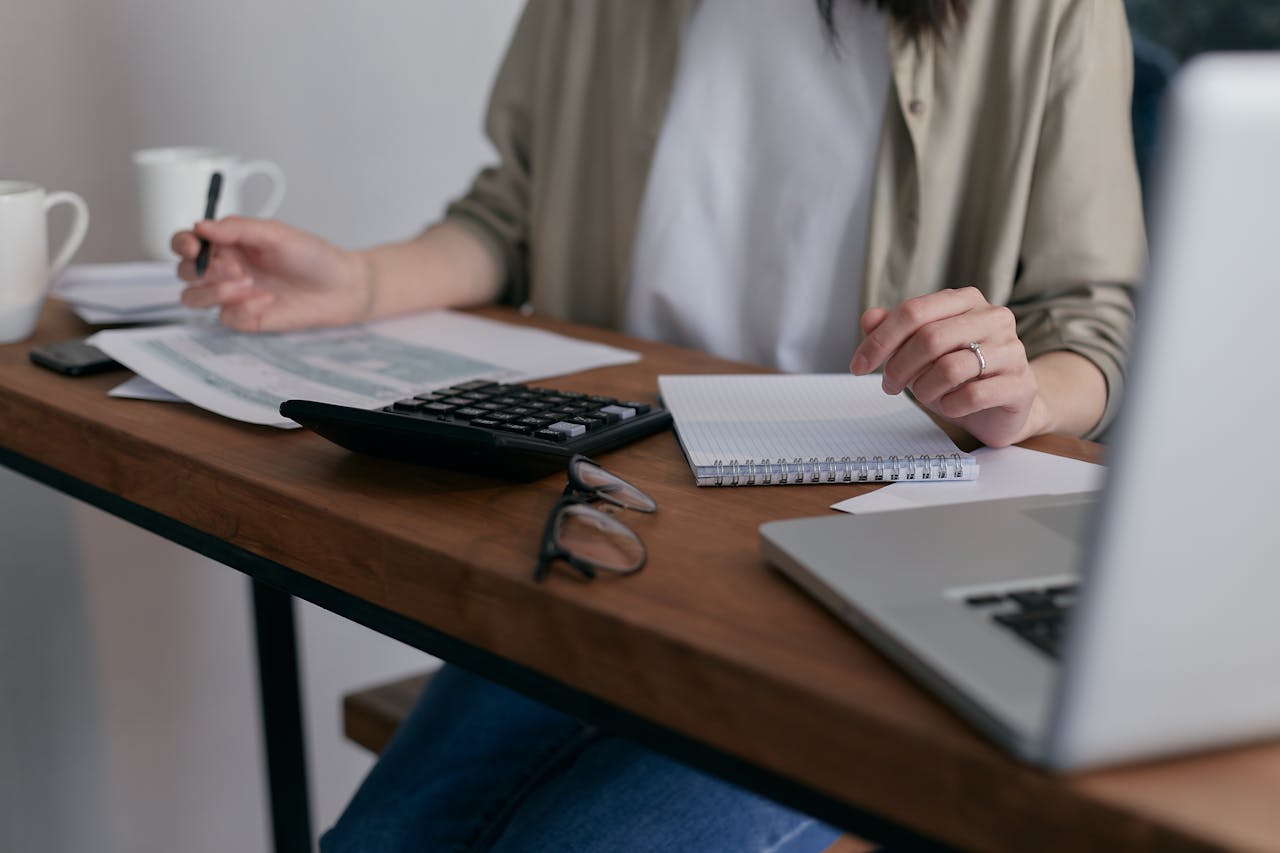 about-us A woman manages finances at home, using a laptop and calculator on a wooden desk.